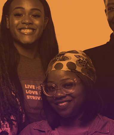 Two people, one standing and one seated, smile at the camera against an orange background. The person standing wears a "Live Here Love Strive" shirt; the seated person wears glasses and a headscarf.