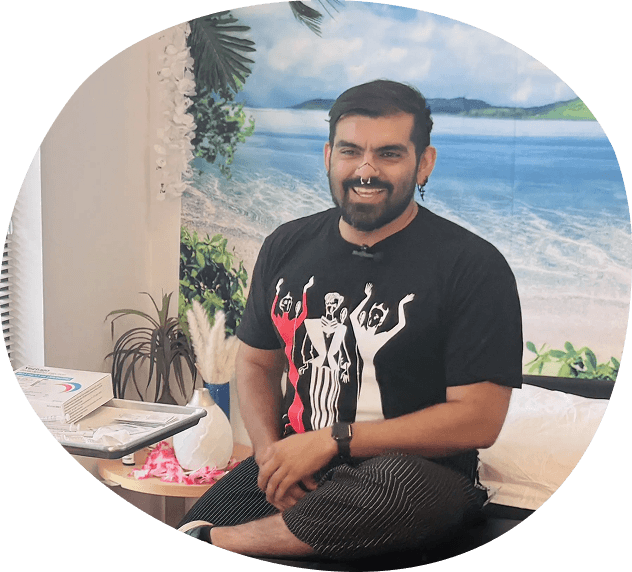 A man with a beard sits cross-legged on a bench, smiling, in front of a beach-themed backdrop. Beside him, a table with plants, books, and items adds to the cozy scene—reminiscent of the welcoming atmosphere at the Whitman Walker Prep Clinic in Washington DC.