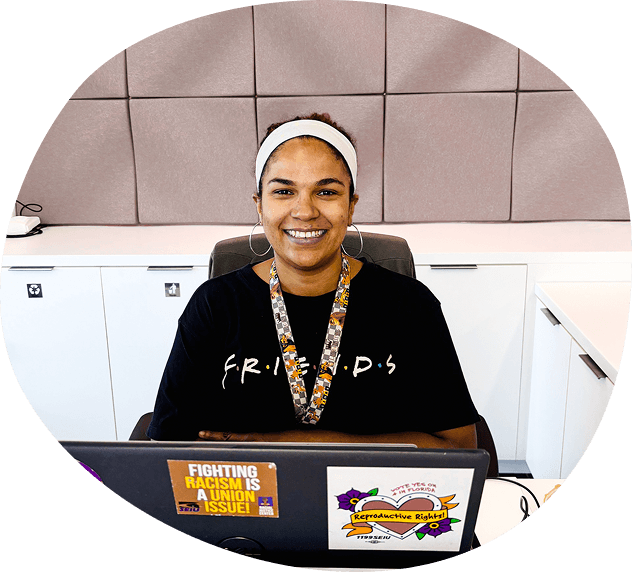 A person sits at a desk with a laptop, wearing a "FRIENDS" shirt and headband, smiling. The laptop has stickers promoting fighting racism and reproductive rights—perfect for someone passionate about social justice issues, such as health care navigators in Washington DC.