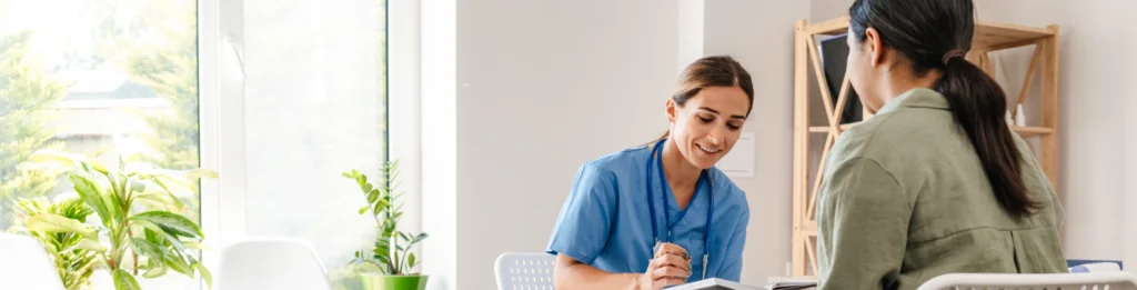 A healthcare professional in blue scrubs sits at a desk, discussing a PEP appointment in Washington DC with a patient in a green jacket in a bright, well-lit office.