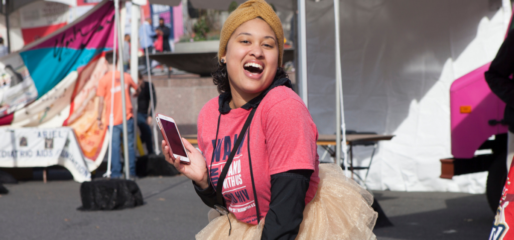 Person wearing a red shirt and tan headband smiles while holding a smartphone at an outdoor event, with tents and banners in the background.