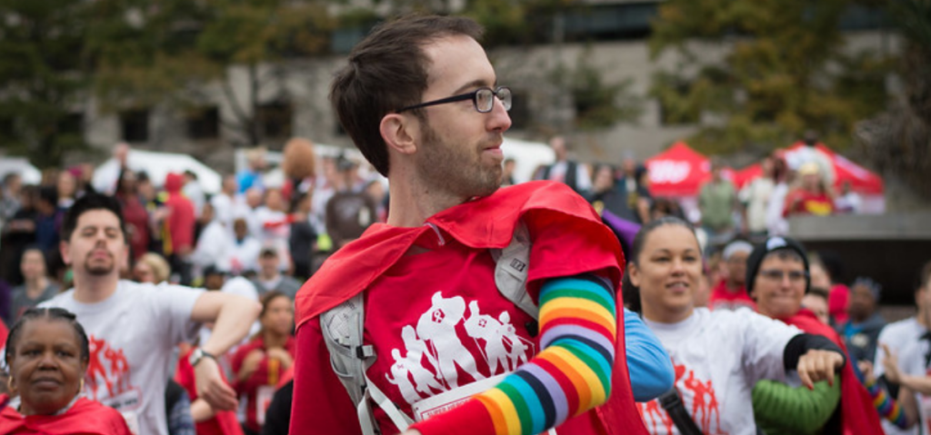 A group of people wearing red and white shirts, with one person in front wearing a rainbow-striped sleeve and red cape, participating in an outdoor event.