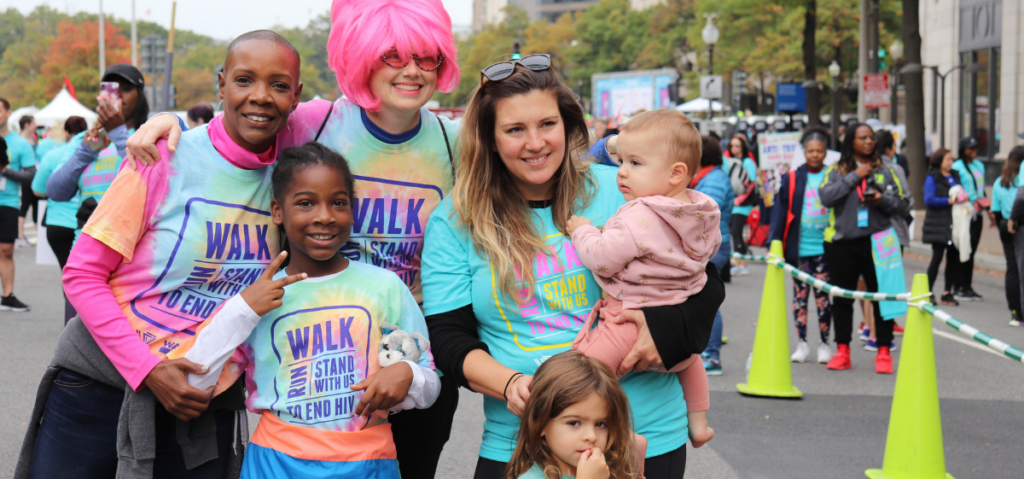 A group of adults and children wearing “Walk to End HIV” shirts pose together at an outdoor event, with other participants in the background.