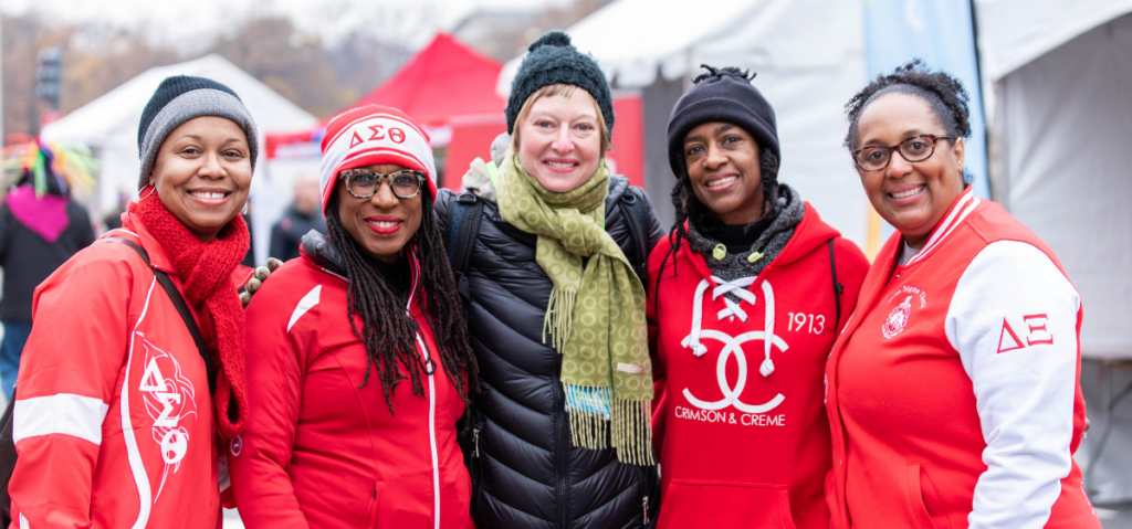 Five women stand together outdoors in winter clothing, smiling at the camera; three wear matching red jackets with Greek letters and symbols, and tents are visible in the background.