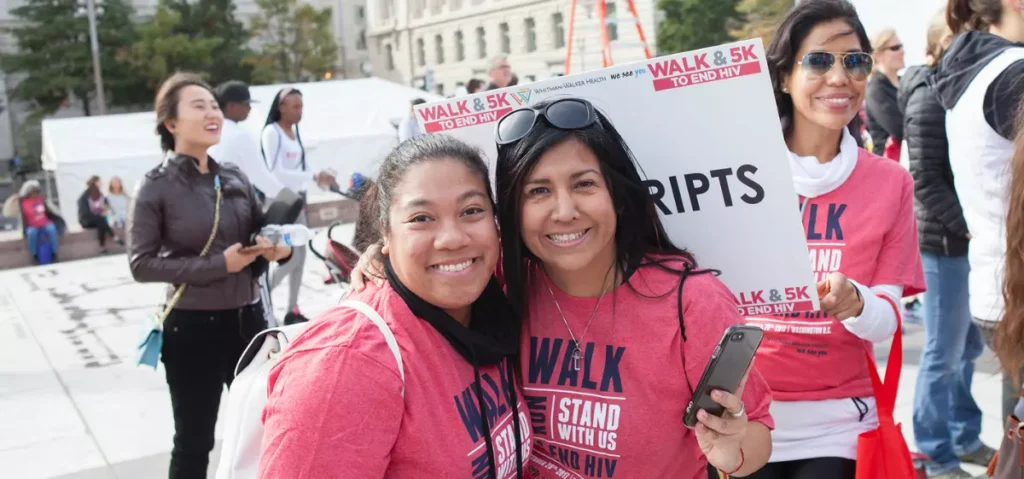 Two women wearing red “Walk & 5K to End HIV” shirts smile at the camera during an outdoor event, surrounded by other participants.