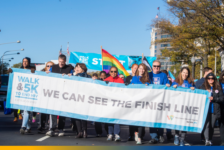 A group of people participate in a walk and 5K event to end HIV, holding a banner that reads “We Can See the Finish Line.”.