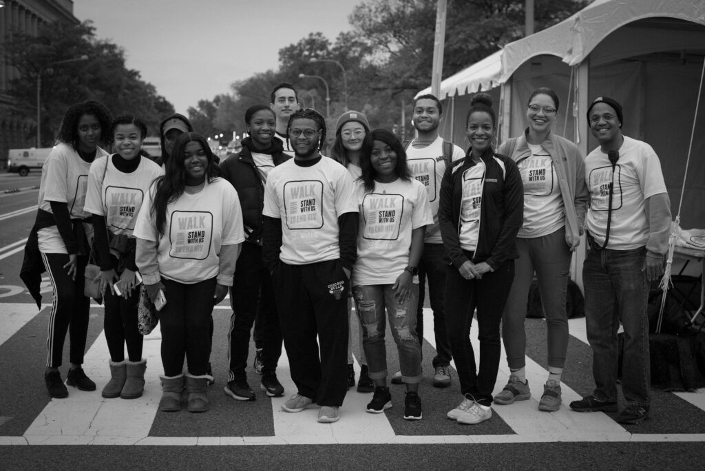 A group of people wearing matching "WALK to SHAW" shirts stand together and pose for a photo at an outdoor event near a tent.
