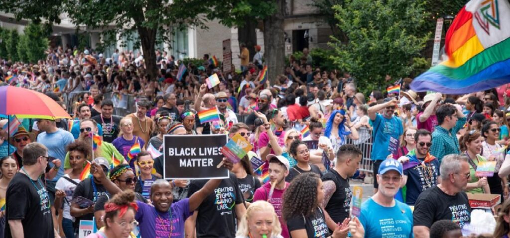 A large crowd participates in a Pride parade, holding rainbow flags and a "Black Lives Matter" sign, with people standing behind barriers along the street.