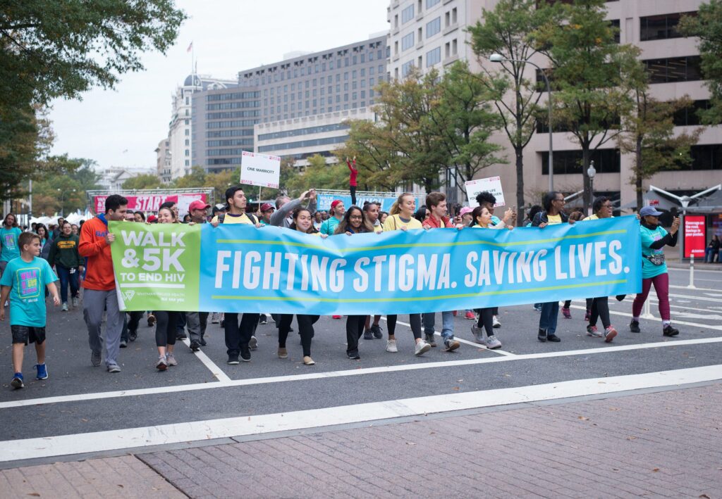 A group of people walk in a city street holding banners that read "Fighting Stigma. Saving Lives." and "Walk & 5K to End HIV.