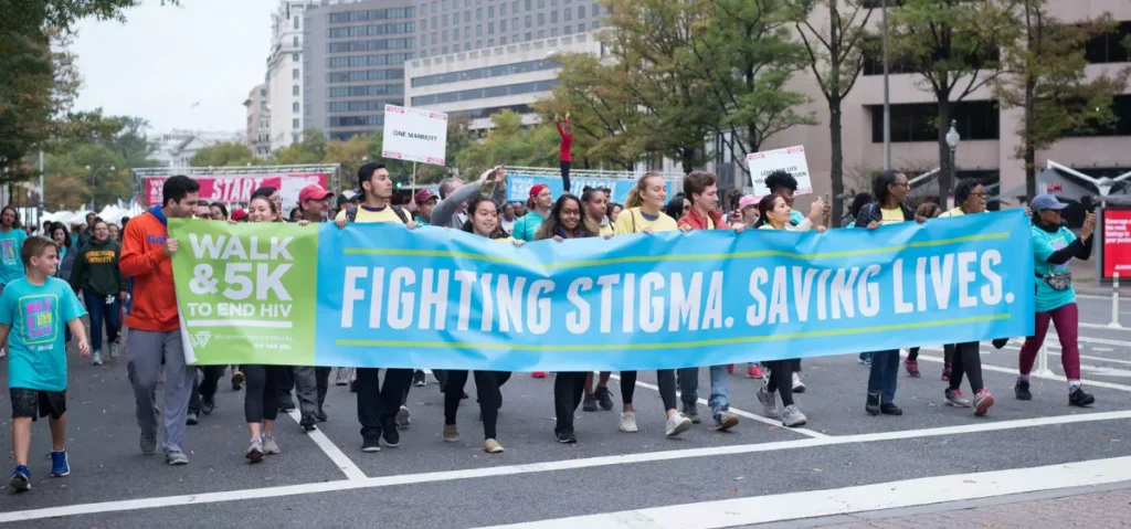 A group of people walk in a city street holding a large banner that reads "Fighting Stigma. Saving Lives." at a Walk & 5K to End HIV event.