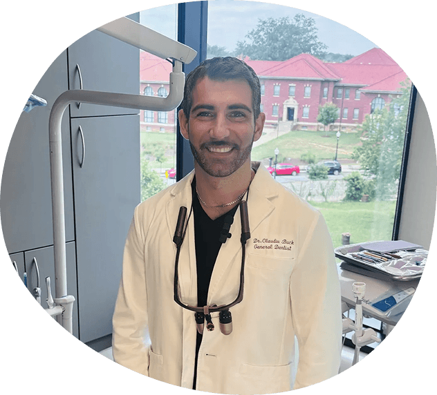 A Washington DC Dentist in a white coat stands in a dental office, smiling, with dental equipment and a window showing buildings and trees in the background.