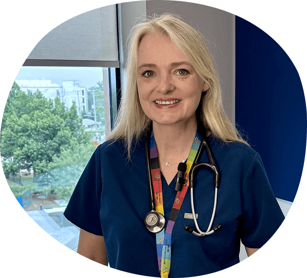 A primary care doctor in Washington DC, dressed in blue medical scrubs with a rainbow lanyard and stethoscope, stands indoors near a window, smiling at the camera.