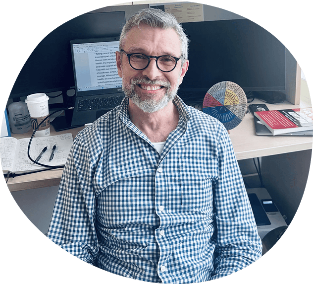 A man with glasses and a beard smiles at a desk with computers, papers, a coffee cup, pens, a color wheel, and books in the background, reflecting the organized yet dynamic environment often found in Behavioral Health DC offices.