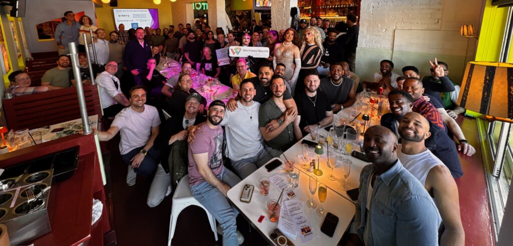 A large group of people gathered around tables in a restaurant, smiling at the camera with drinks and menus visible on the tables during a lively drag brunch event.