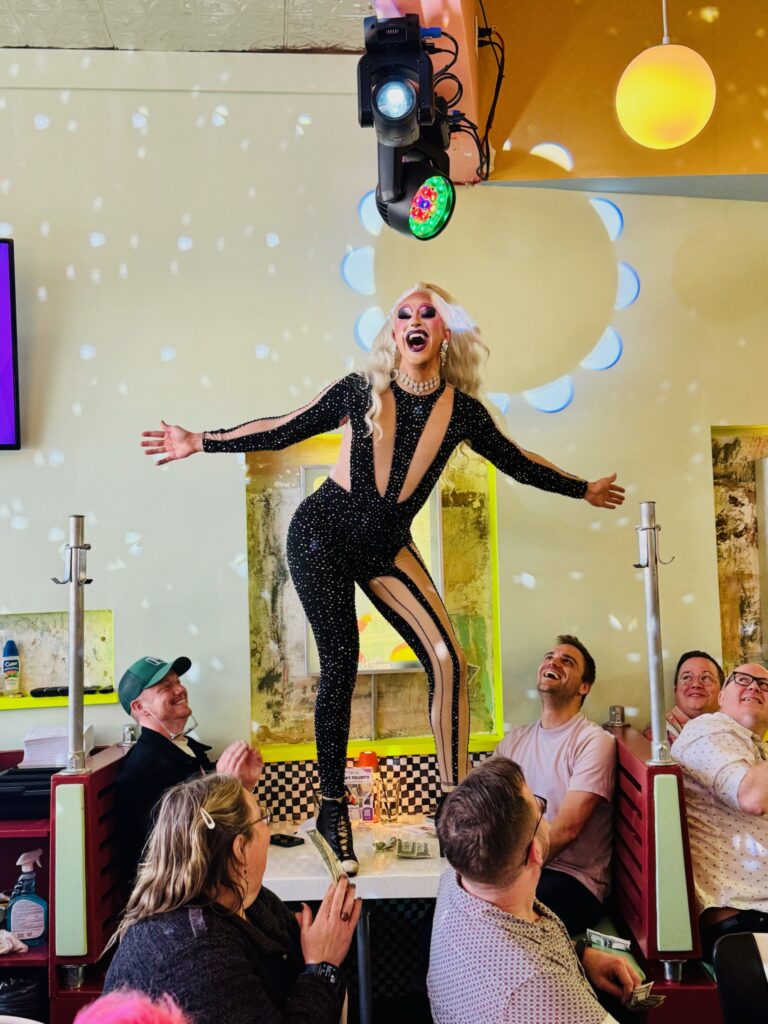 A drag performer in a black, sparkling bodysuit poses confidently on top of a booth, while an audience seated around cheers and smiles.