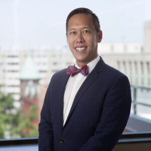 A man in a navy suit and red bow tie stands indoors in front of a window overlooking a cityscape, representing Whitman Walker Legal Services with professionalism.