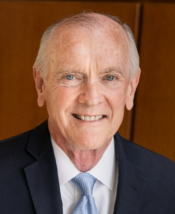 An older man in a dark suit, light blue tie, and white shirt smiles at the camera in front of a wooden background, representing Whitman Walker Legal Services with warmth and professionalism.