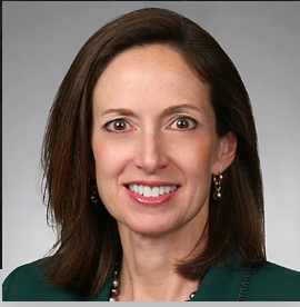A woman with straight brown hair and brown eyes, wearing a green blazer and pearl earrings, smiles at the camera against a gray background, representing Whitman Walker Legal Services.