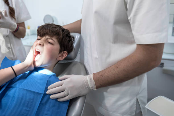A boy sits in a dental chair holding his cheek in pain while a dentist in white gloves stands beside him.