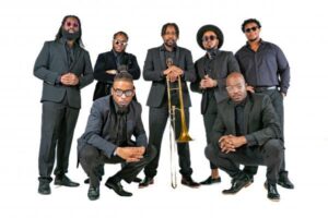 Seven men dressed in black suits pose together against a white background, with one holding a trombone in the center.