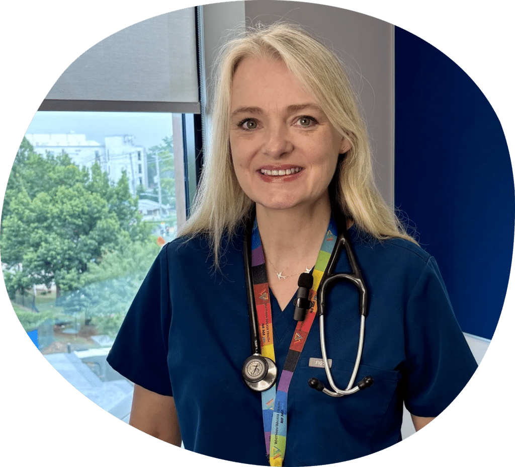 A woman in blue scrubs with a stethoscope around her neck stands indoors by a window, smiling at the camera.