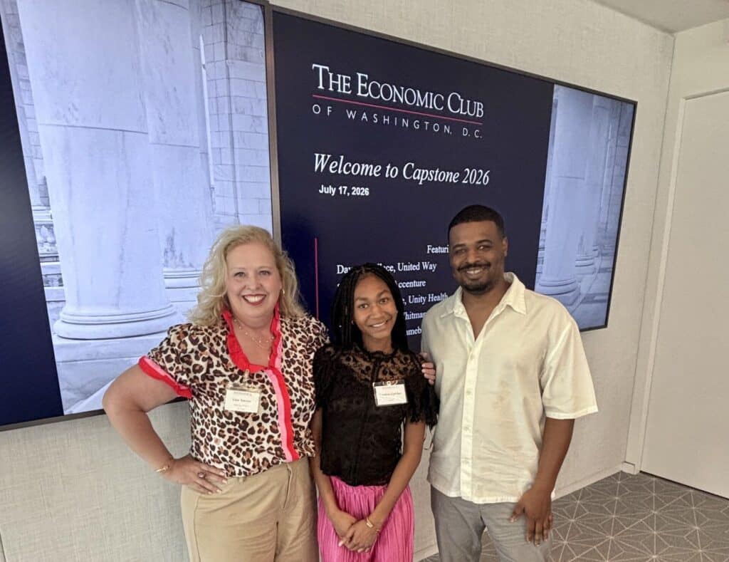 Three people stand and smile in front of a sign reading "The Economic Club of Washington D.C. Welcome to Capstone 2026." The sign includes the date July 17, 2026.