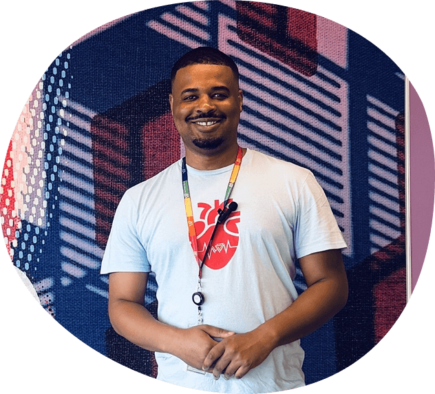 A man wearing a light blue Whitman Walker shirt with a heart graphic stands in front of a colorful, geometric-patterned wall, smiling at the camera.