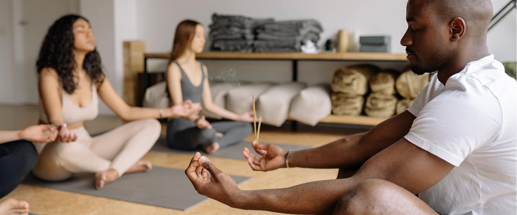 Four people sit cross-legged on yoga mats meditating in a studio, with incense burning and rolled towels and mats stacked on shelves in the background.