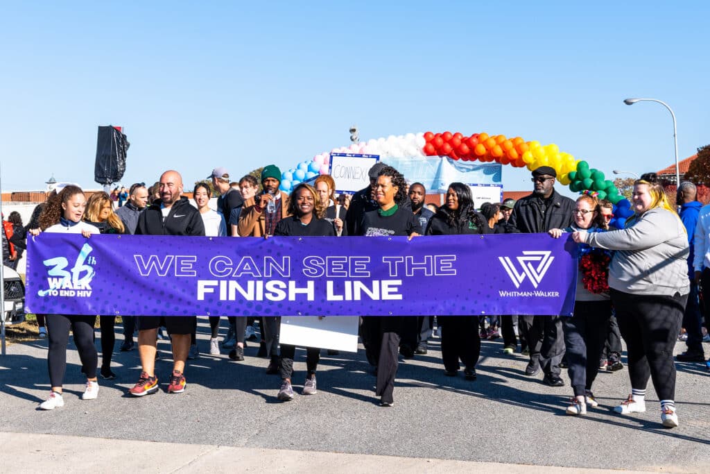 A diverse group of people walk outdoors, holding a “We Can See The Finish Line” banner at an organized event. A rainbow balloon arch is visible in the background, highlighting the enthusiastic spirit of the Walk to End HIV which aims to raise funds for Whitman-Walker.