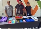 Three people stand behind a table displaying brochures, purple tote bags, and bottled water at a community health event.