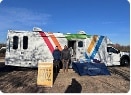 Two people stand in front of a recreational vehicle decorated with colorful diagonal stripes, with a folding sign and table set up on the ground.