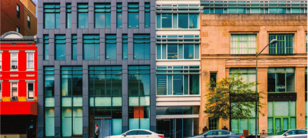 Three modern and historic buildings stand side by side on a city street with parked cars and a few pedestrians walking by.