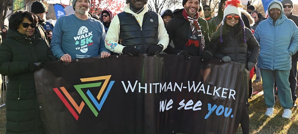 A group of people outdoors hold a "Whitman-Walker: We see you." banner at a Walk to End HIV event; others stand in the background on a sunny day, capturing the spirit seen in walk to end hiv event photos.