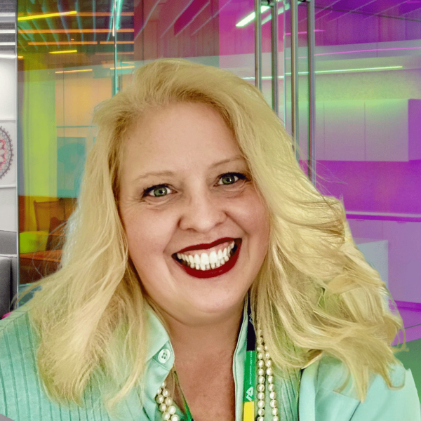 A woman with blonde hair and a wide smile, wearing a light green shirt and pearl necklace, is in a colorful, modern office setting with glass walls and neon lighting.