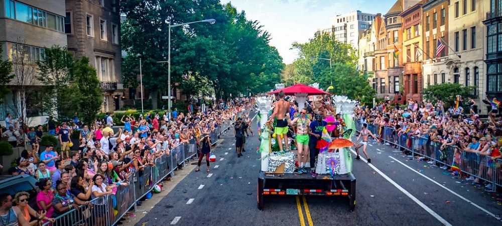 A parade float with people in colorful outfits moves down a city street lined with cheering crowds behind barriers on a sunny day.