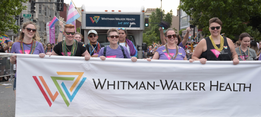 A group of people march in a parade holding a large Whitman-Walker Health banner, with a truck and pride flags visible in the background.