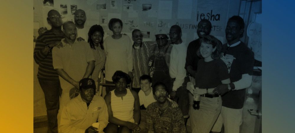 A group of people pose together indoors in front of a wall covered with papers and posters. Some are standing, while others are seated or kneeling in the front row.