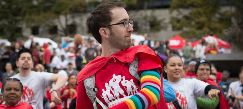 A group of people wearing red and white shirts, with one person in front wearing a rainbow-striped sleeve and red cape, participating in an outdoor event.