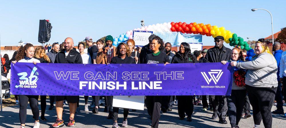 A diverse group of people walk outdoors, holding a “We Can See The Finish Line” banner at an organized event. A rainbow balloon arch is visible in the background, highlighting the enthusiastic spirit of the Walk to End HIV which aims to raise funds for Whitman-Walker.