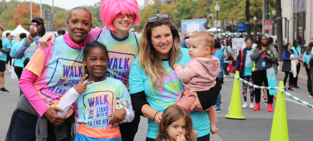 A group of adults and children wearing “Walk to End HIV” shirts pose together at an outdoor event, with other participants in the background.