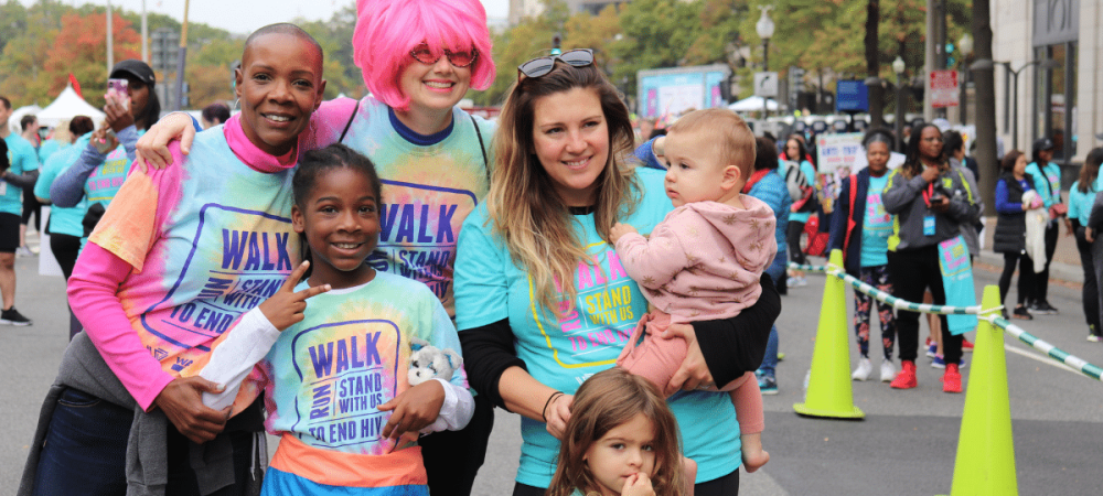 A group of adults and children wearing “Walk to End HIV” shirts pose together at an outdoor event, with other participants in the background.