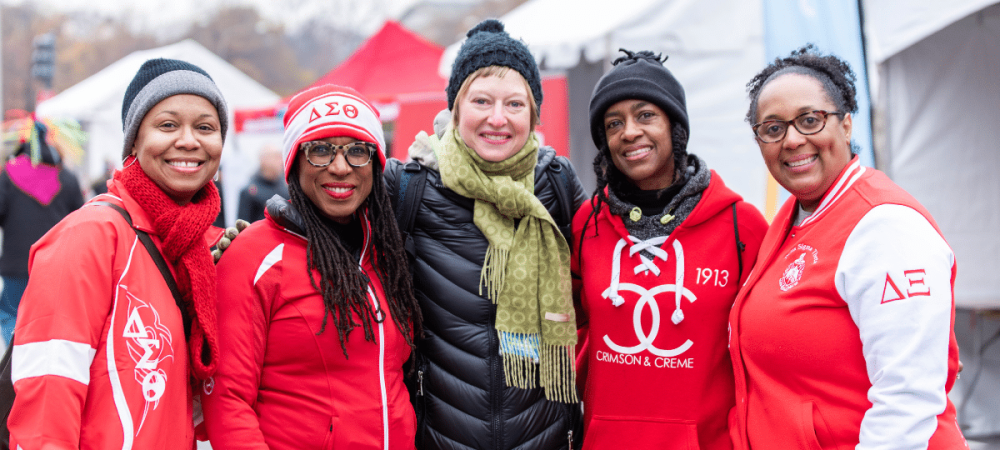 Five women stand together outdoors in winter clothing, smiling at the camera; three wear matching red jackets with Greek letters and symbols, and tents are visible in the background.