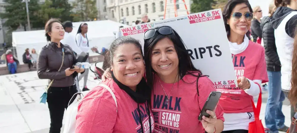 Two women wearing red “Walk & 5K to End HIV” shirts smile at the camera during an outdoor event, surrounded by other participants.