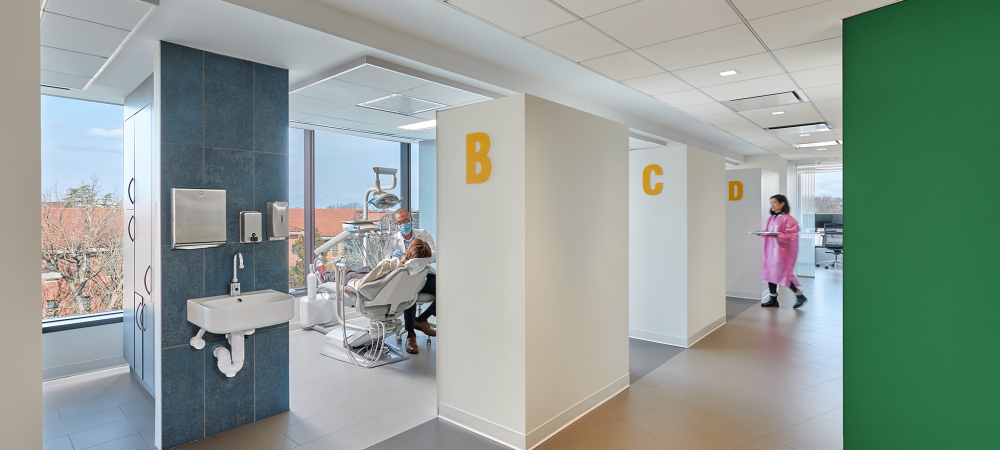 A dental clinic with four exam rooms labeled A to D; during the Max Robinson Open House, a patient is seated in room B as a staff member in pink scrubs walks down the hallway.