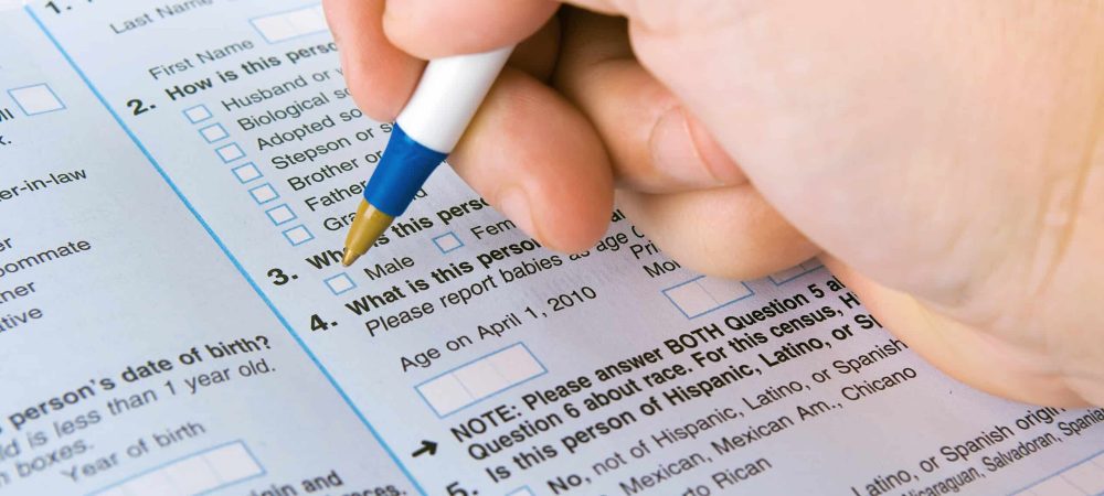 A hand holding a pen fills out a census survey form with checkboxes and questions about gender, age, ethnicity, and LGBTQ questions.