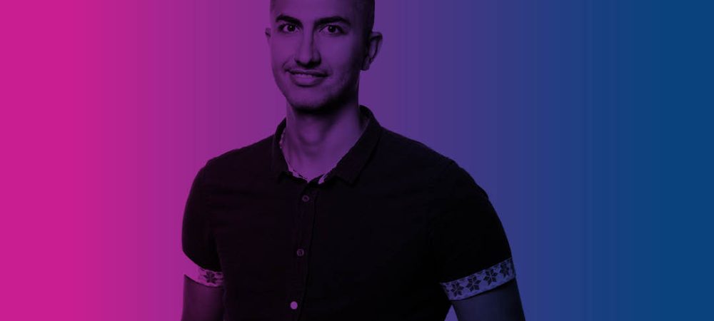 A person stands facing forward with a neutral expression, set against a gradient background transitioning from pink on the left to blue on the right.