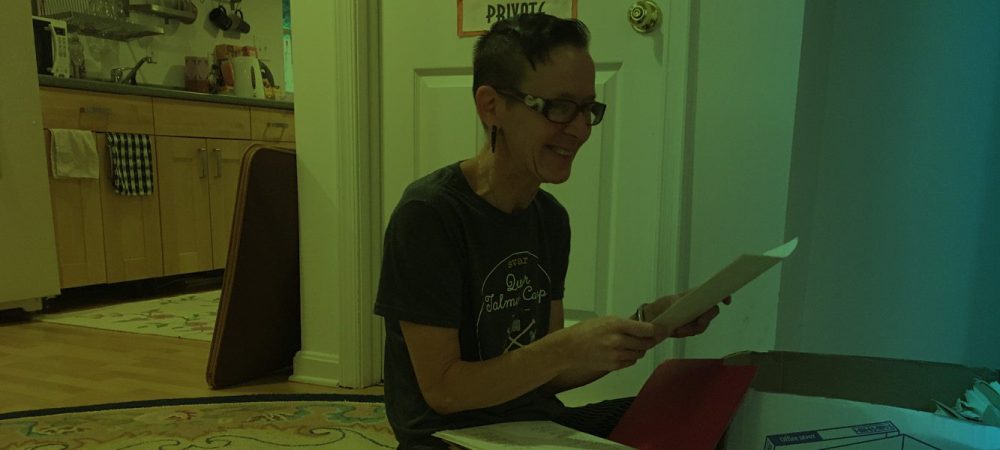 A person sitting on the floor reads papers from a folder, with a box nearby. A kitchen and a door with a "PRIVATE" sign are in the background.