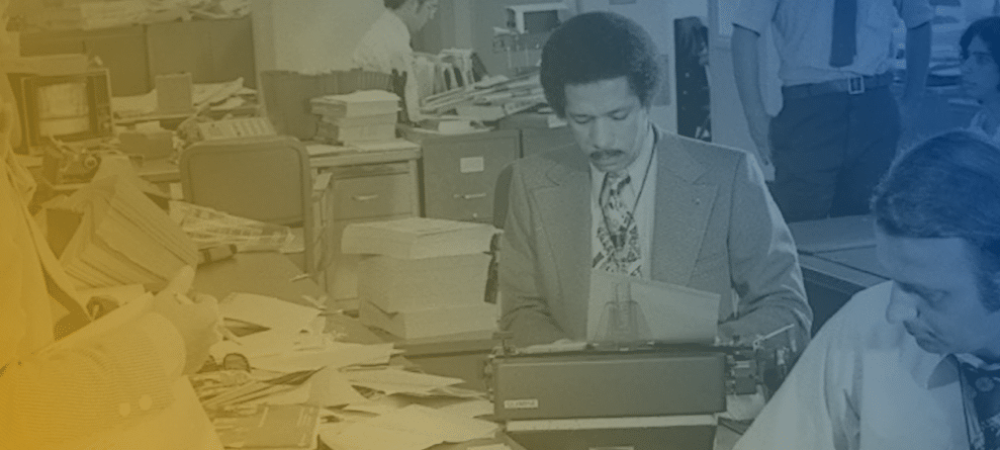 Several people work at cluttered desks in a busy office, with one man typing on a typewriter in the foreground.