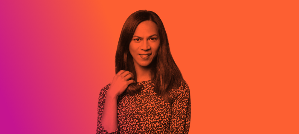 Person with long hair wearing a patterned top stands against a vibrant orange and pink gradient background, looking at the camera.