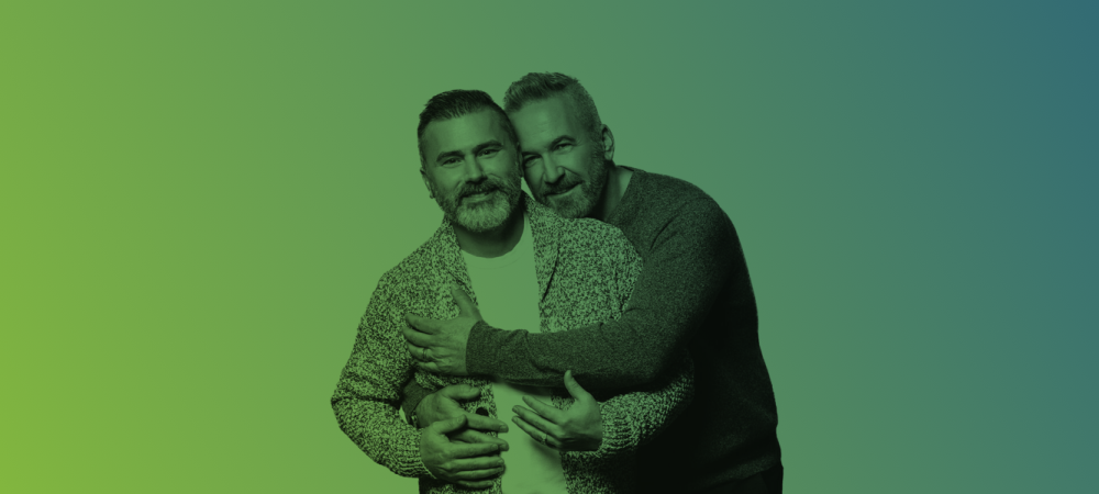 Two men with beards standing close together, one standing behind and hugging the other, both facing the camera against a green to blue gradient background.